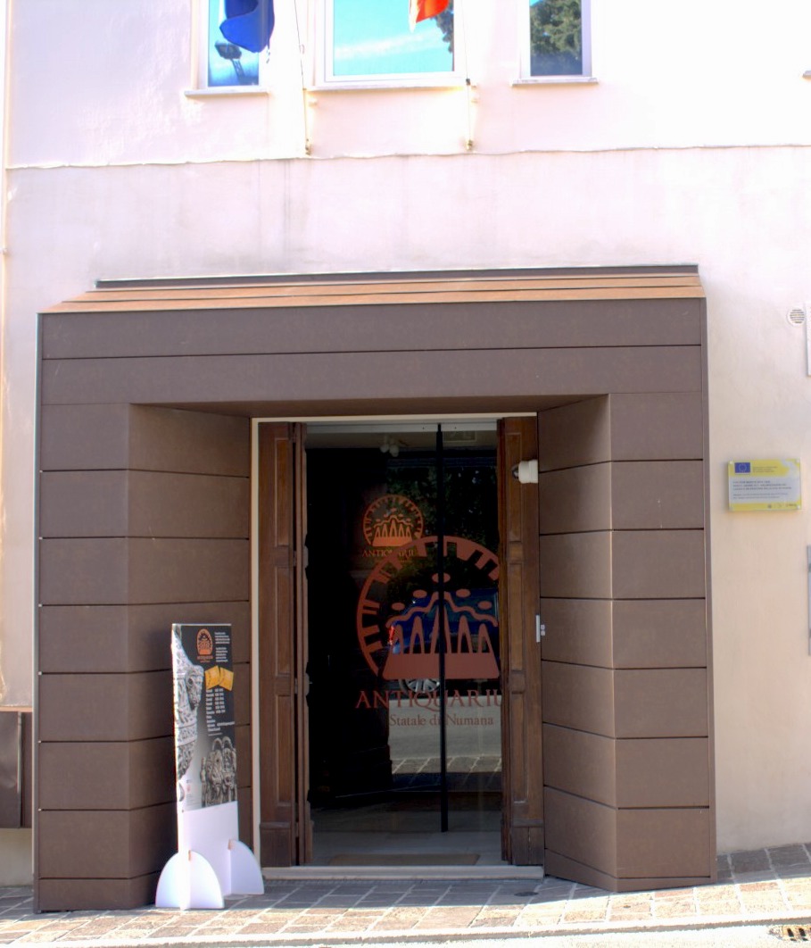 Vista frontale dell'ingresso dell'Antiquarium statale di Numana, che si apre sul piano stradale. Il varco della porta d'ingresso, spicca sulla superficie dell'edificio perché incassata in una spessa cornice aggettante le cui pareti interne sono disposte oblique a formare come un largo imbuto e invitano a varcare la soglia. Sulla soglia della porta è presente un pannello promozionale del museo, su piedistallo a terra. Sulla parete esterna dell'efidicio, a destra della spessa cornice, è affissa la targa ufficiale che identifica il museo. (Foto: E. Giovagnoli)