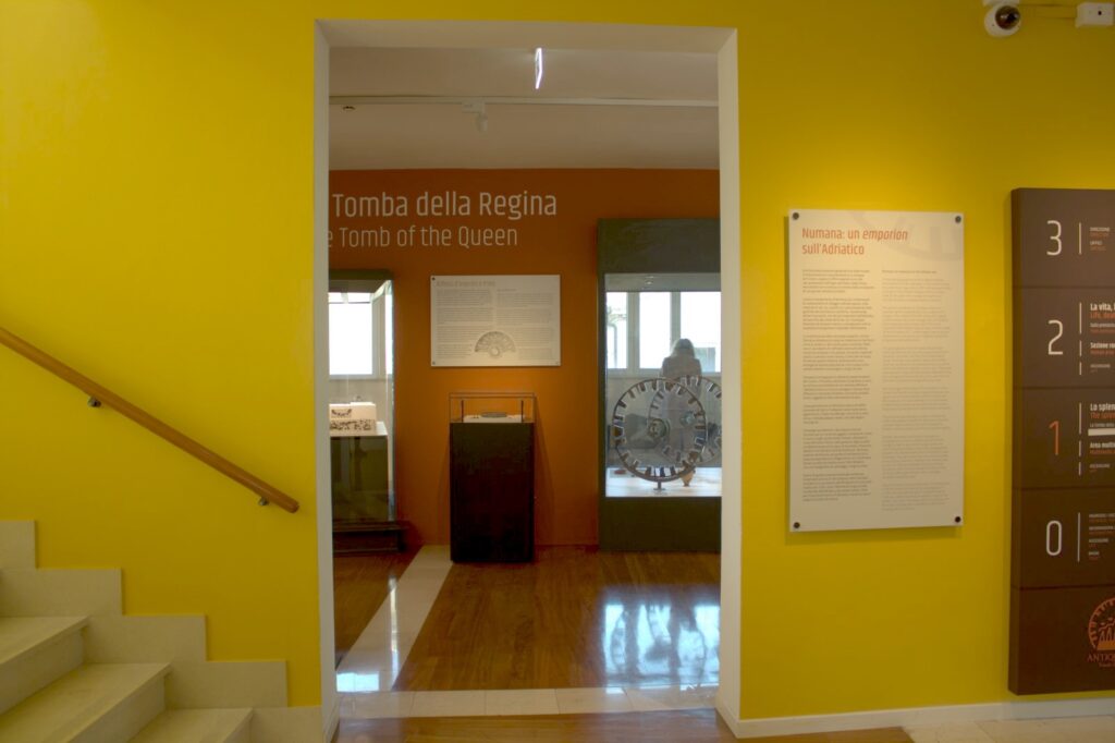 Inquadratura dell'interno del museo, di fronte a una grande porta senza ante che si apre nel corridoio principale del museo, al di là si scorge parte delle vetrine dell'allestimento dello scavo archeologico della «Tomba della Regina». Immediatamente a destra della grande porta, sulla parete, è affisso un pannello esplicativo e ancora più a destra un pannello della segnaletica di orientamento del museo che indica i piani e le sale. (Foto: E. Giovagnoli)